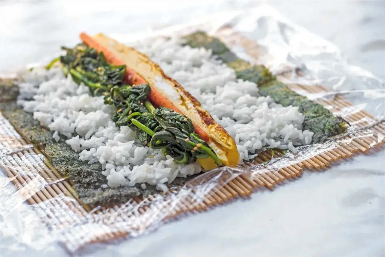 Przygotowanie domowego sushi: ryż, nori, szpinak i omlet na macie bambusowej.